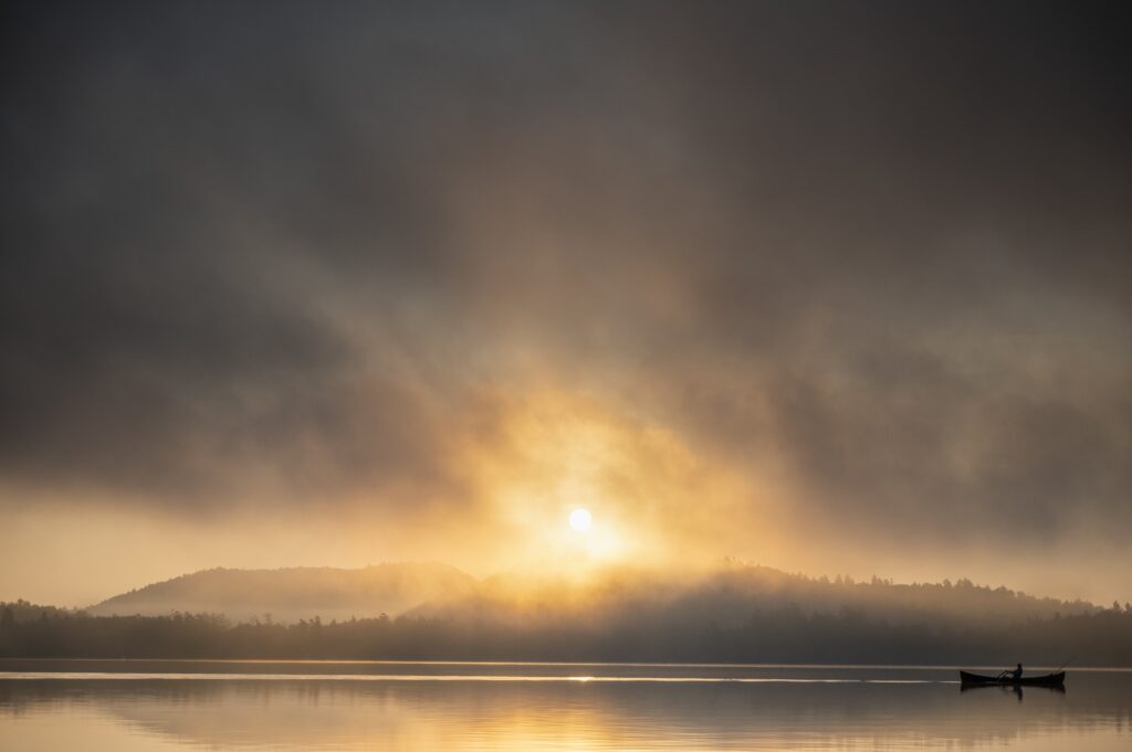A boater rows their wooden boat in the distance as the sun rises above a lake