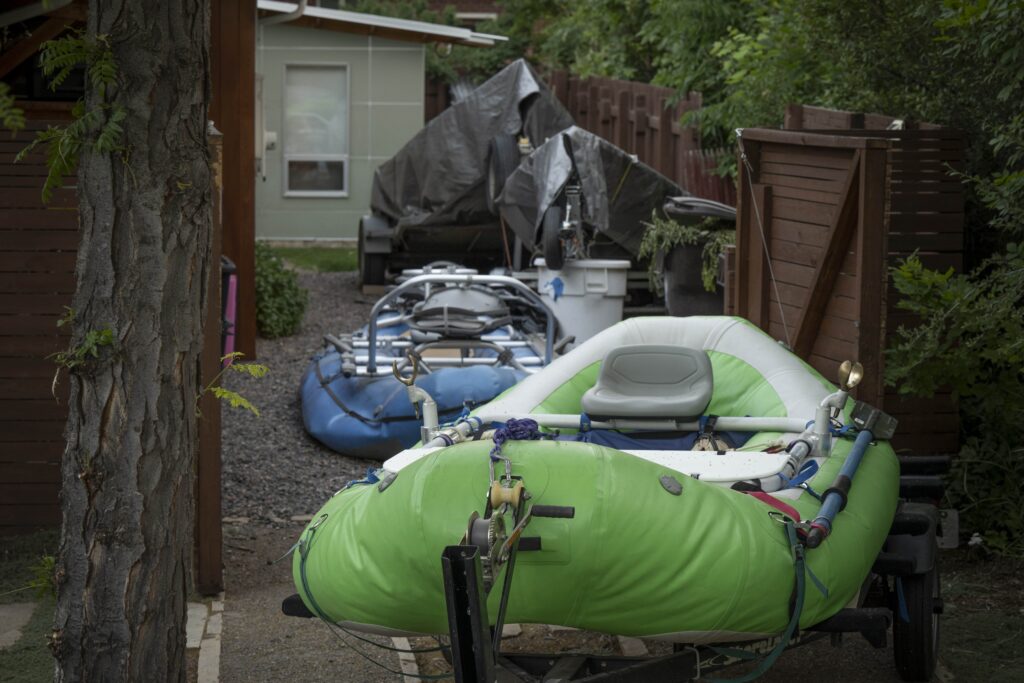 Two fishing rafts, one green and one blue, sit on trailers in a gravel driveway next to a house.