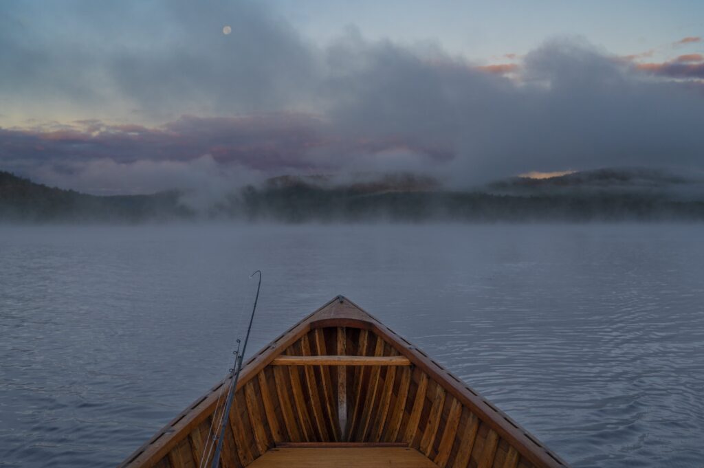 A bent fishing rod hangs over a wooden boat amid morning fog on a lake