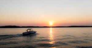 A speedboat navigates a serene lake as the sun sets, casting colorful reflections on the water.
